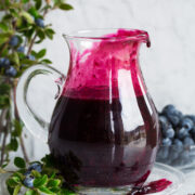 Blueberry syrup in a glass pitcher set over a glass plate.