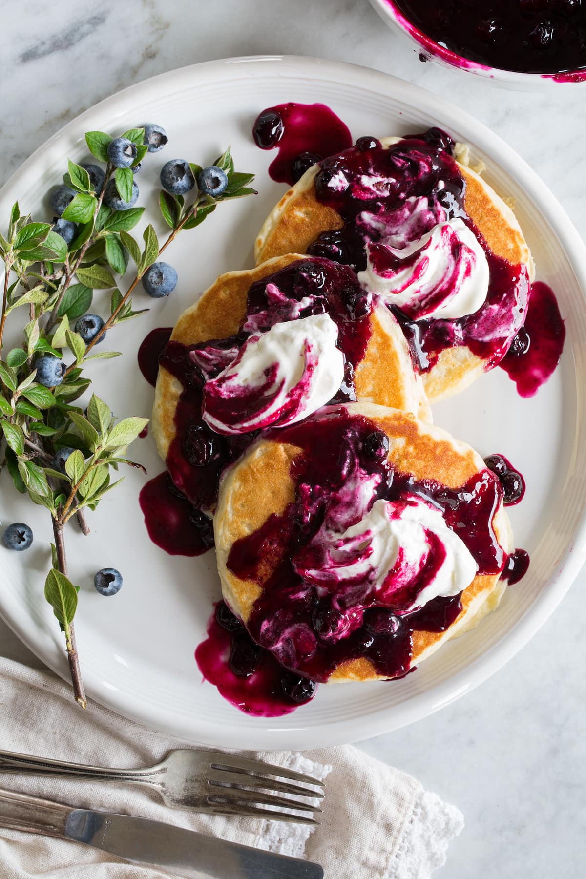 Blueberry syrup shown poured over pancakes with whipped cream on top.