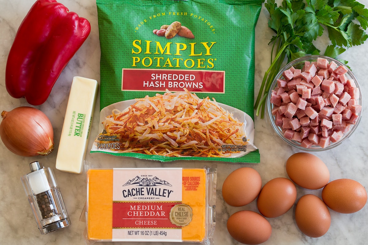 Photo: Ingredients shown that are used to make breakfast hash. Includes packaged shredded hash browns, bell pepper, cheddar cheese, eggs, butter, onion, ham, parsley, salt and pepper.