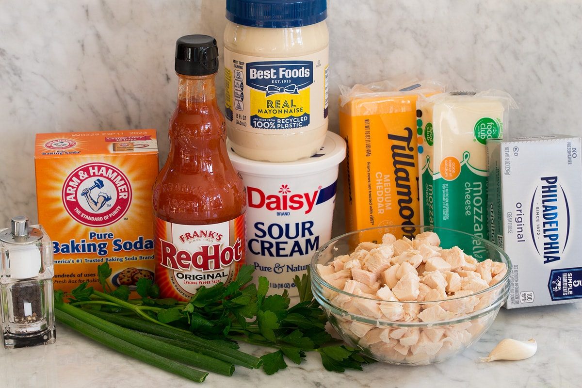 Photos of ingredients for buffalo chicken dip. Includes cooked chicken, buffalo sauce, sour cream, mayonnaise, cheddar cheese, mozzarella cheese, cream cheese, garlic, green onions, parsley, baking soda, salt and pepper.
