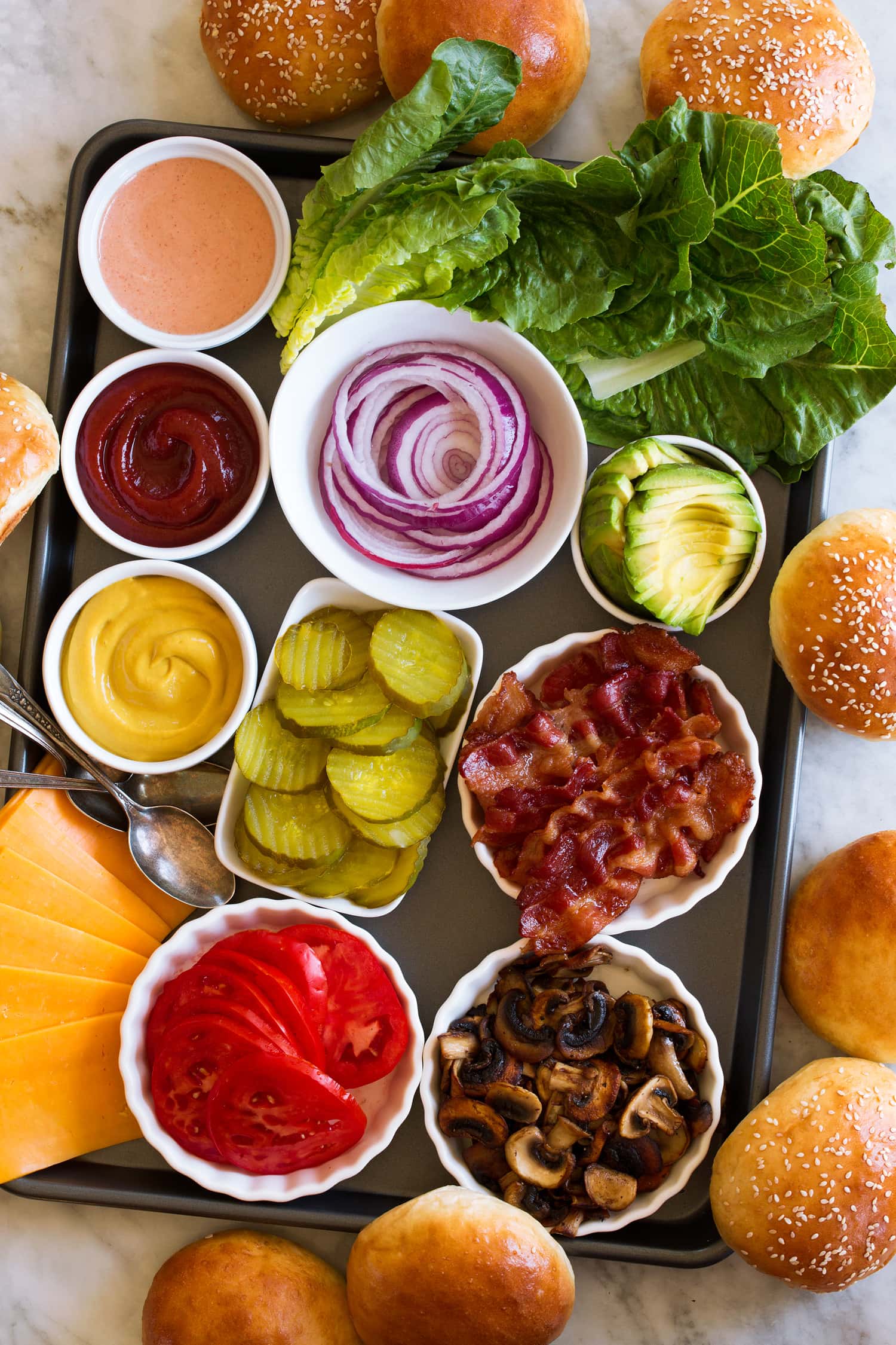 Topping ideas for hamburgers shown on a baking sheet with hamburger buns surrounding them.