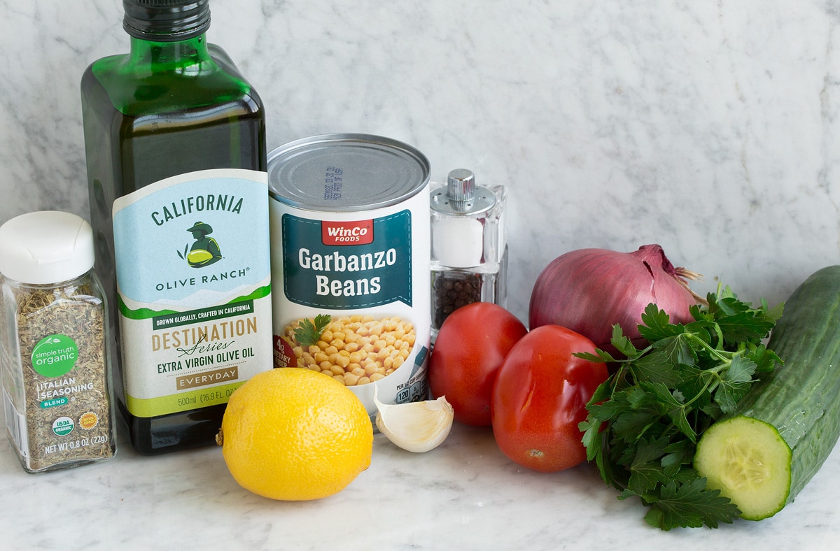 Photo of ingredients used to make chick pea salad. Includes olive oil, chickpeas/garbanzo beans, tomatoes, red onion, parsley, cucumber, garlic and lemon.