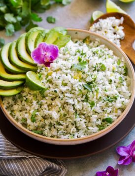Serving bowl full of cilantro lime rice garnished with avocado slices, cilantro leaves, orchid flower and lime.