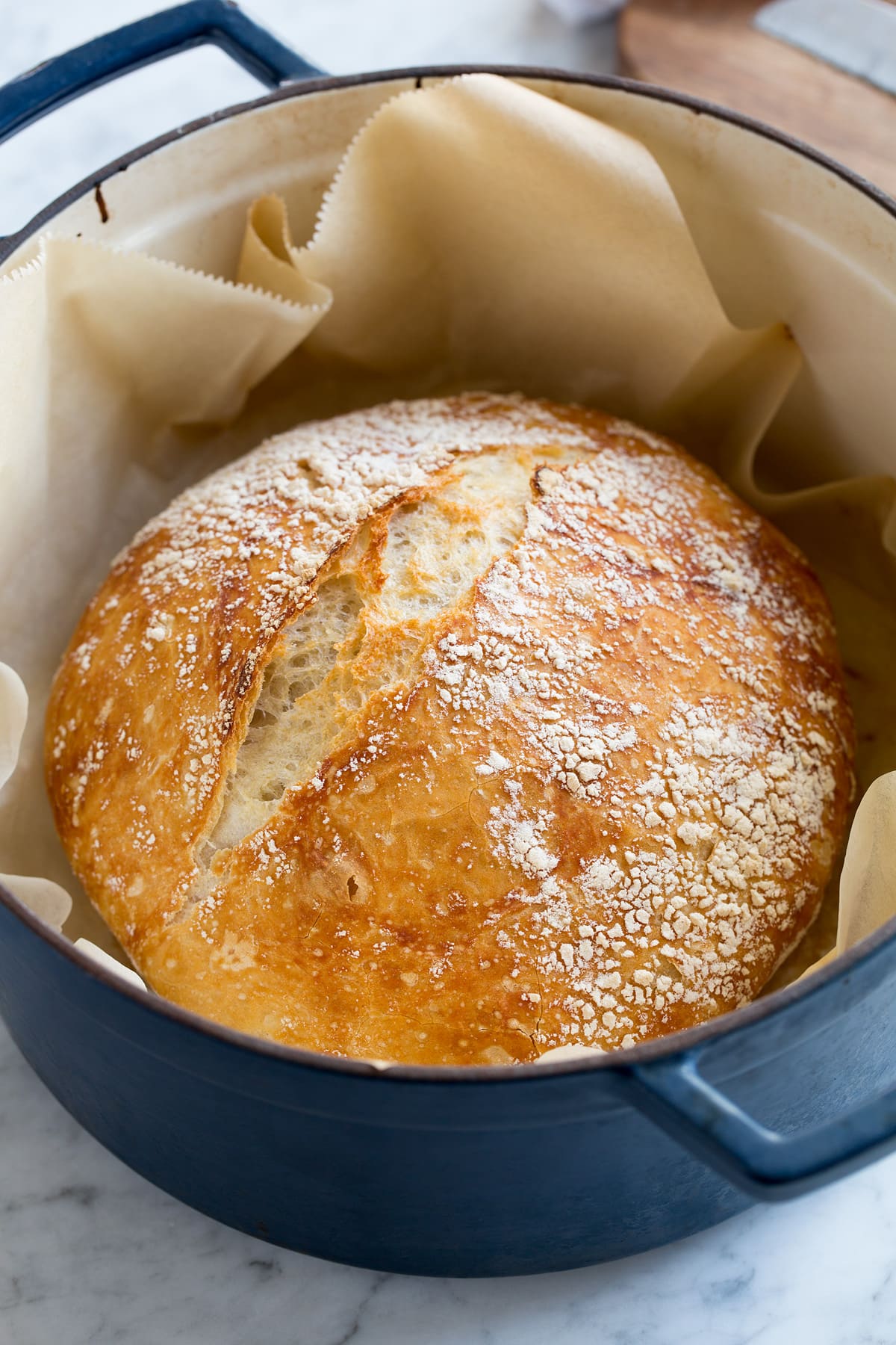 No knead bread in a large blue enameled cast iron pot. Bread is sitting on a sheet of parchment paper and pot is resting on a marble surface.