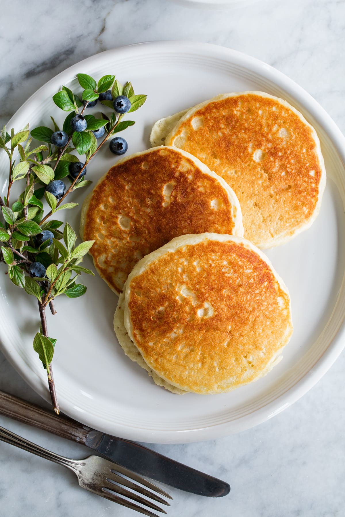 Three pancakes spread out on a large white plate with a blueberry stem to the side.