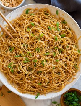 Photo: Bowl of sesame noodles topped with peanuts, sliced green onions and sesame seeds. Shown overhead in a white bowl with a dark napkin underneath and chopsticks and serving spoons to the side.