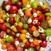 Tomato Salad in a white serving bowl. Shown in salad are multi-color cherry tomatoes and grape tomatoes, mozzarella pearls and it's coated in a herb dressing.