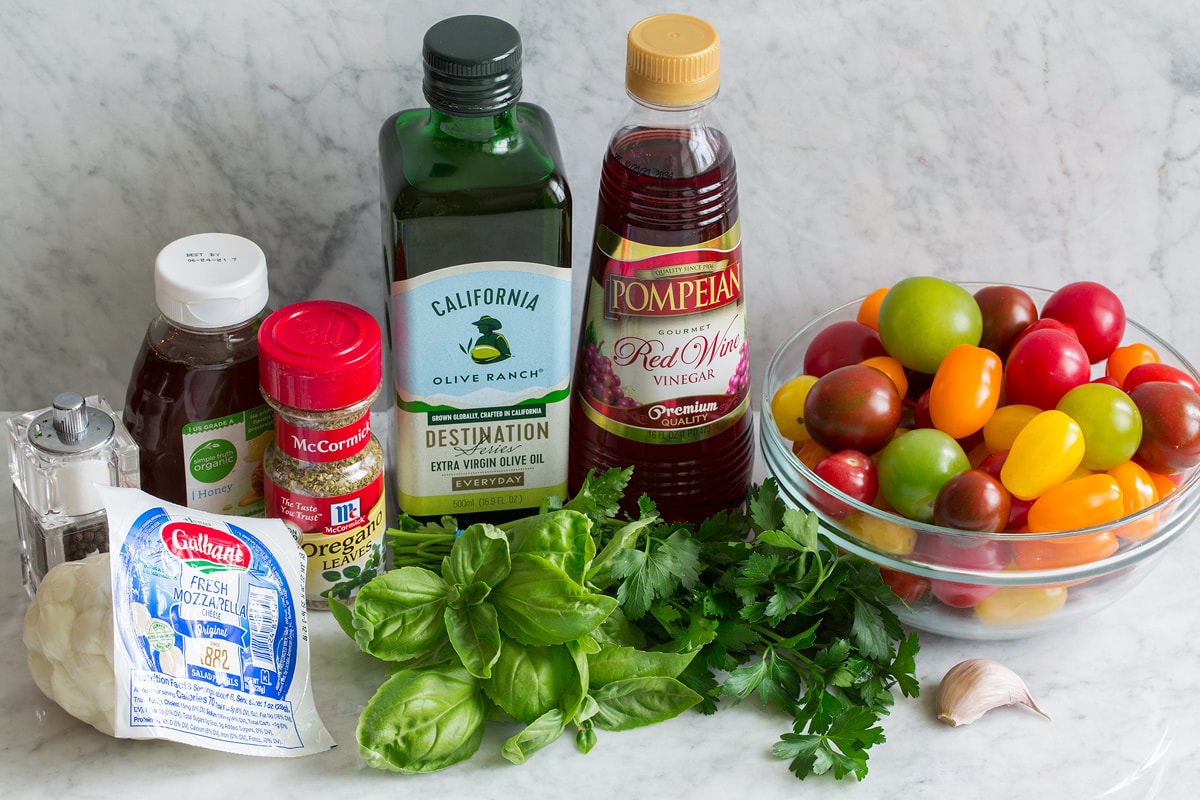 Image of ingredients used to make tomato salad. Includes cherry and grape tomatoes, fresh basil and parsley, garlic, fresh mozzarella, dried oregano, honey, olive oil, red wine vinegar and salt and pepper.