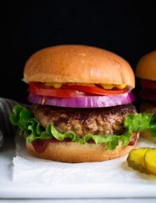 Turkey Burger layered with bun, ketchup, lettuce, turkey patty, red onion, tomato and yellow mustard. Served with a side of pickles on a white platter.