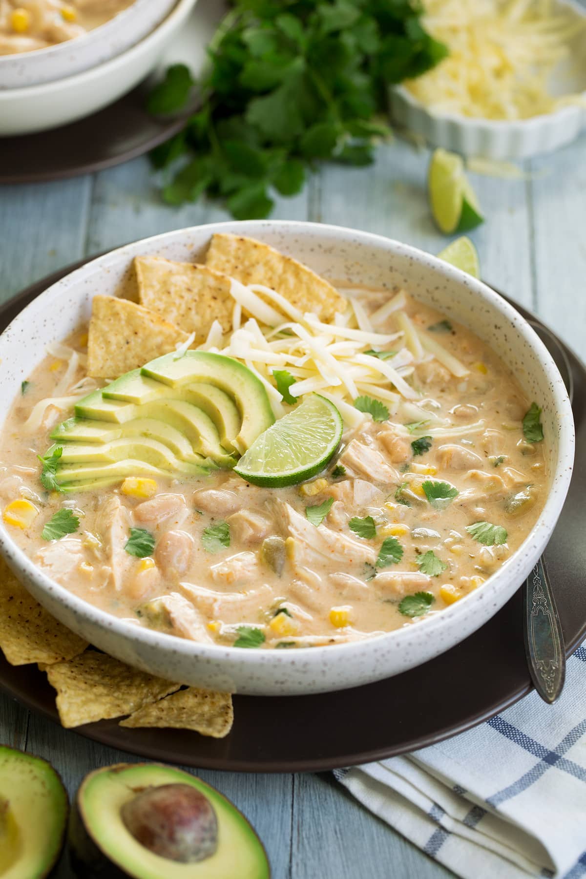 White Chicken Chili in white serving bowl set over a brown plate. Chili is garnished with avocado, cheese, tortilla chips, cilantro and lime.