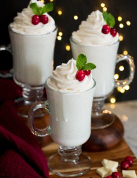 Three mugs of white hot chocolate on a wooden tray with Christmas lights in the background.